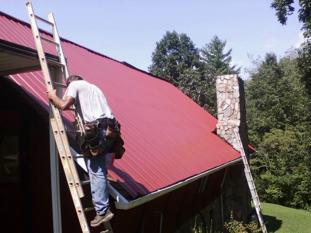 Roofer on a ladder beside a red metal roof and stone chimney — Soffit & Fascia Repair by Roofing Brothers in Needville